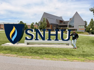 Saptarsi Mondal wearing a conference badge beside the large SNHU sign on the Southern New Hampshire University campus, with a modern gabled building behind.