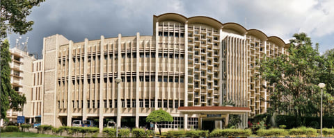 The main building of IIT Bombay, a long modernist facade with rows of vertical fins, behind trees and a lawn.