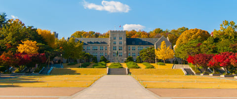 The stone main hall of Korea University at the top of a wide lawn and stairway, framed by trees in red and gold autumn colours.